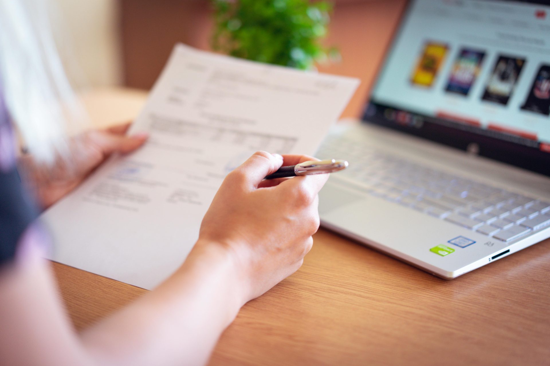Nahaufnahme einer Hand, die Stift und Papier neben einem Laptop hält – ein Bild, das Online-Shopping oder die Durchsicht von Dokumenten assoziiert.